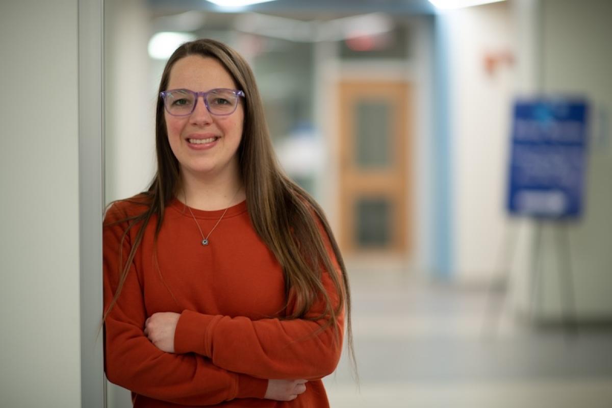 Chelsea Baker, class of 2015, leans against a white doorway. She is wearing an orange long-sleeved shirt and glasses. She's smiling directly at the camera while an out-of-focus hallway is in the background.