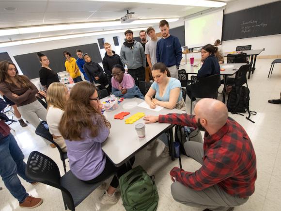 Students gather around a white table with pink and yellow pieces of paper on it. Dr. Moore, in a red and black shirt, crouches next to them.
