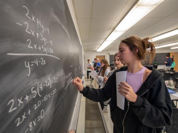 Student wearing black sweatshirt and pink shirt writing math formulas on chalkboard.
