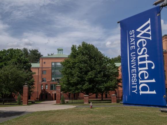 Courtney Hall with blue skies behind it and a Westfield State University blue and white flag in front of it.
