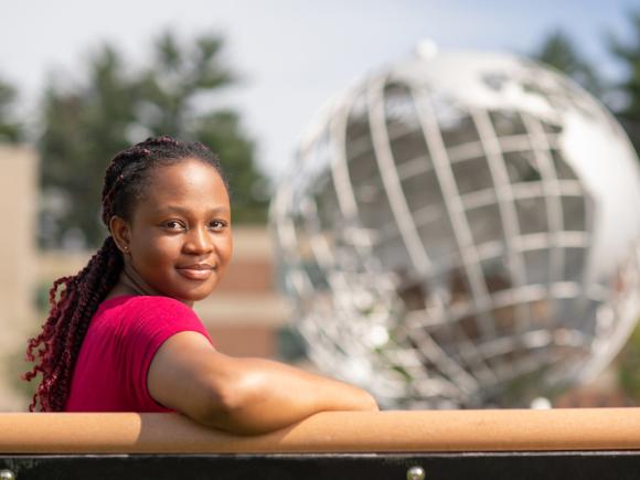 Marilyn Maison, class of 2024. She is sitting outside, on a bench in front of the silver, campus globe. The background behind her is blurred, and she has one arm slunk over the railing she leans against.