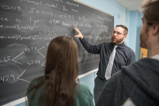 Math instructor in front of chalkboard wearing tie, black shirt, and glasses.