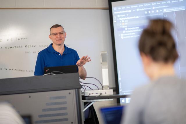 A Computer and Information Sciences professor stands in front of a white board while instructing students in a computer lab.
