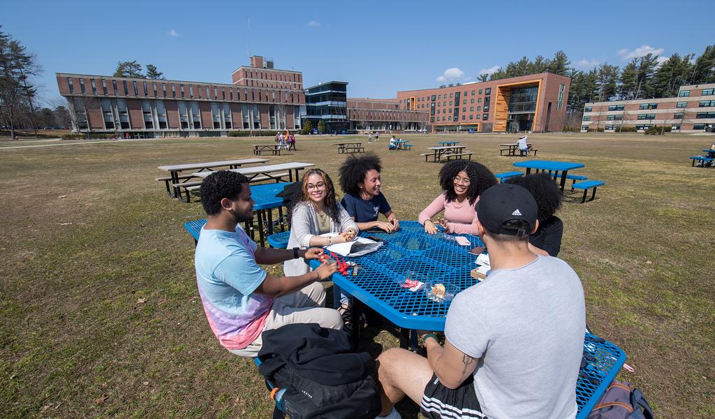 Six students talking and smiling at blue table on campus green with University Hall and Ely Campus Center behind them.