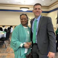 Senator John Velis and an attendee of this year's Chamber of Commerce breakfast. Her name tag is unclear. He is dressed in a suit with a green tie, and she wears a white-and-green sweater along with a sheer, green shawl. Black glasses sit atop her head. In the background, white-clothed tables full of plates and food rests and one of the Colleen's Court women from Holyoke stands off to the side as she talks to someone else.