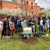 This year's Arbor Day. Students and staff from the biology department gather around a newly planted tree. A white and green sign saying "Arbor Day" is also planted in front of the tree.